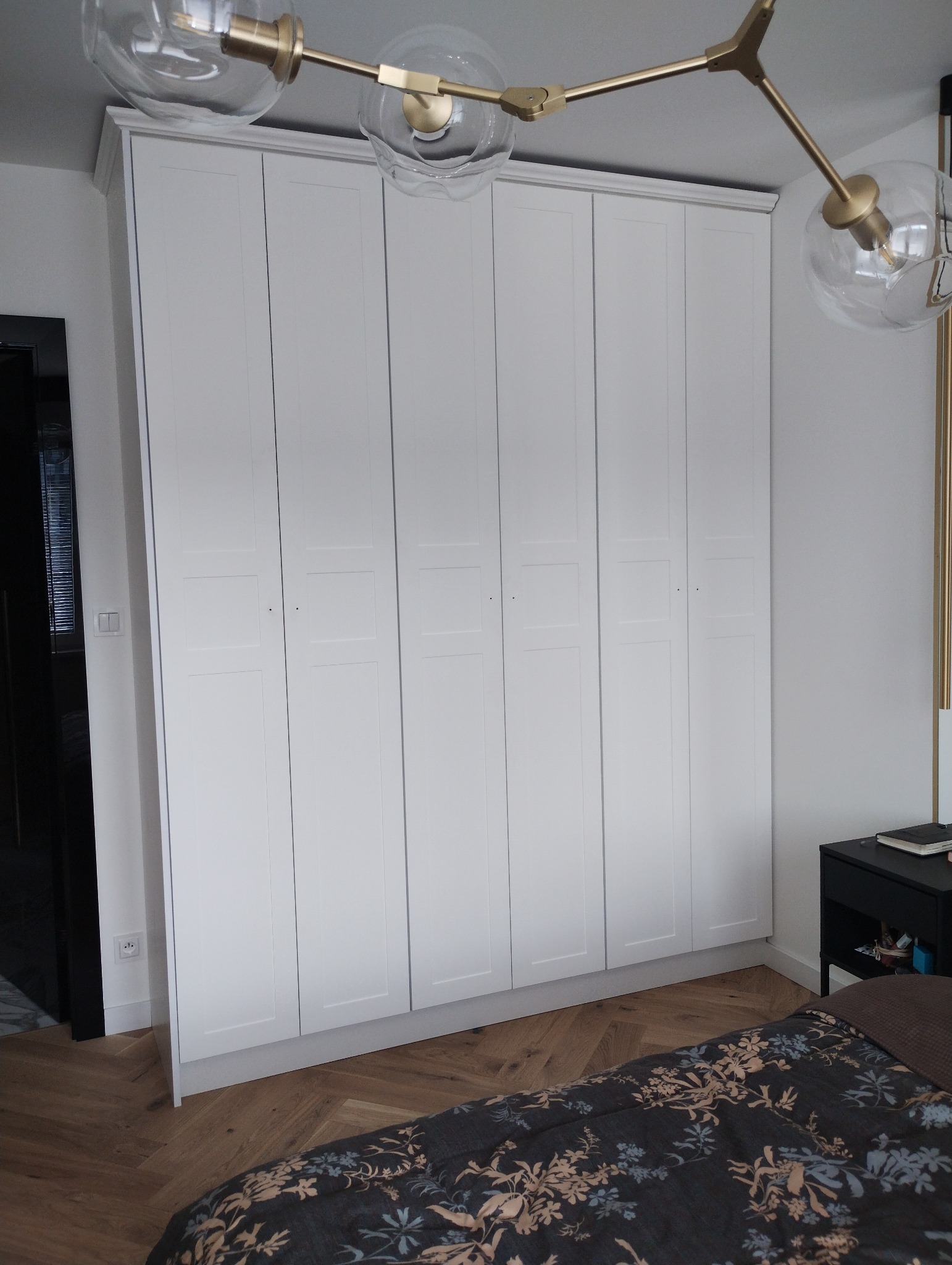 Biała, wysoka szafa wnękowa z sześcioma drzwiami w sypialni, widoczna część łóżka z wzorzystą pościelą i lampa sufitowa z kloszami w kształcie kul.