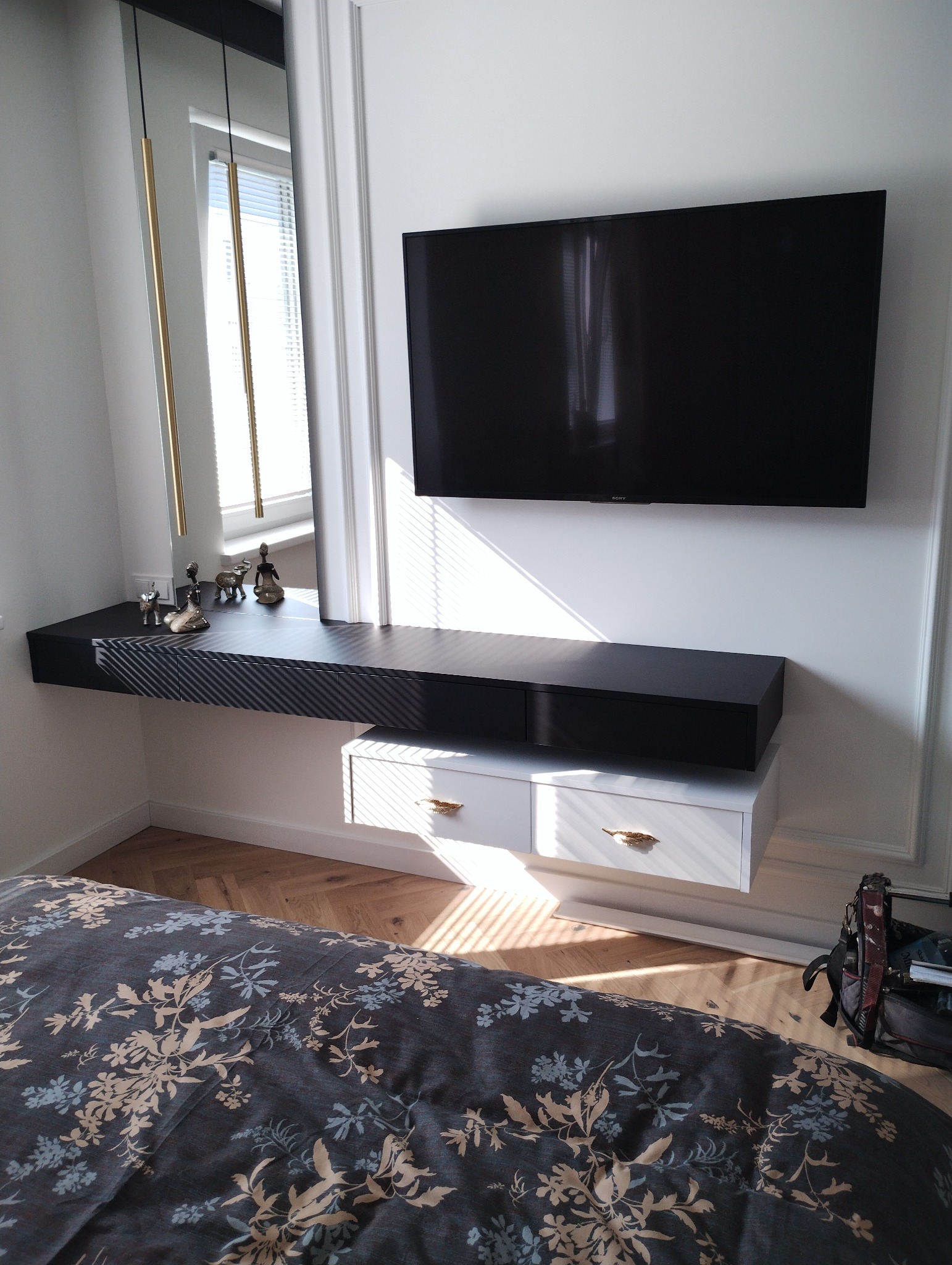 Nowoczesna szafka RTV z lustrem w złotej ramie, czarnym blatem z wzorem i białymi szufladami z ozdobnymi, złotymi uchwytami w kształcie liści, zawieszona pod telewizorem na ścianie z dekoracyjną...