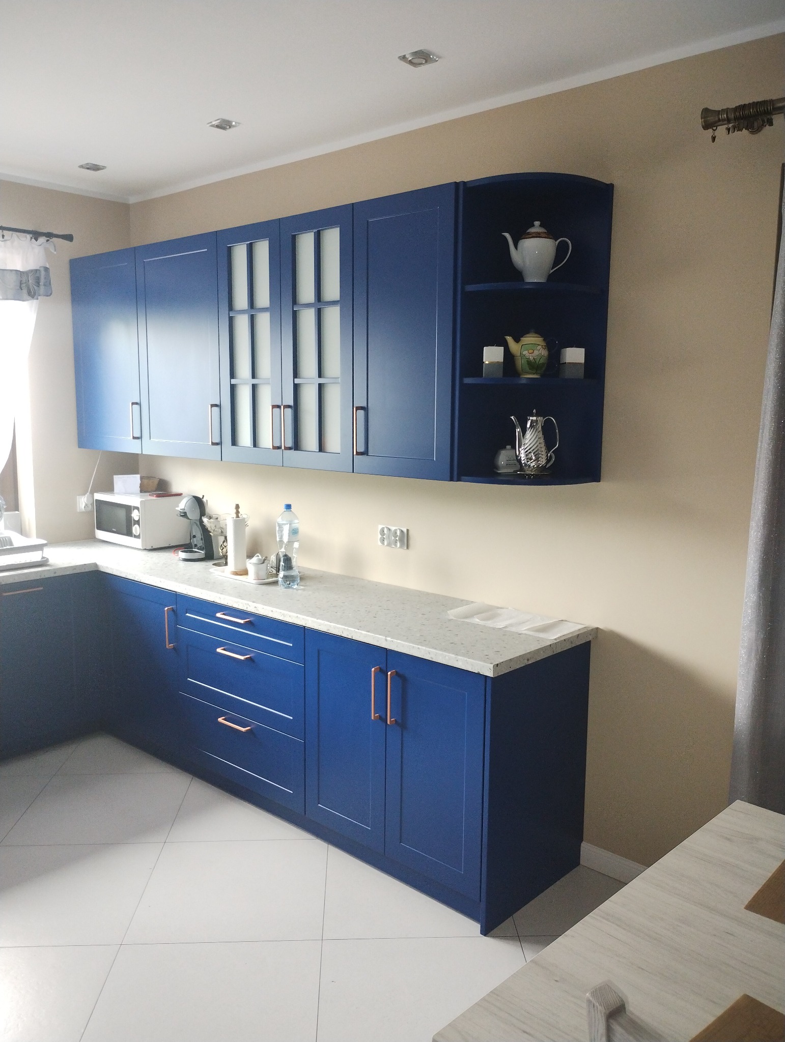 Nowoczesna, granatowa kuchnia z białym blatem i miedzianymi uchwytami, uzupełniona o przeszklone szafki wiszące i otwartą półkę z dekoracyjną ceramiką.