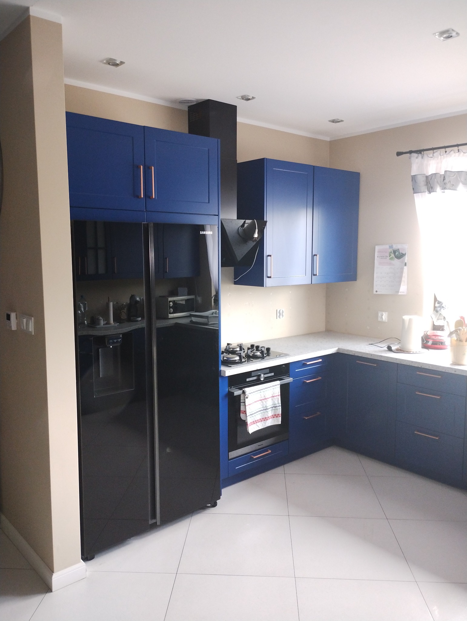 Nowoczesna, granatowa kuchnia z miedzianymi uchwytami, czarną lodówką Samsung i piekarnikiem z zawieszoną ściereczką.