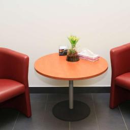 Centrum Zdrowia Psychicznego CZP Tychy - Poczekalnia: Dwa czerwone fotele klubowe po bokach okrągłego stolika z pomarańczowym blatem, na którym stoi szklany wazon z żółtymi kwiatami i pudełko chusteczek.