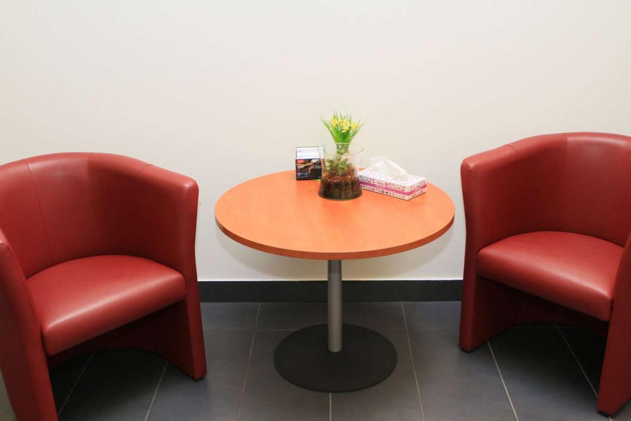 Poczekalnia: Dwa czerwone fotele klubowe po bokach okrągłego stolika z pomarańczowym blatem, na którym stoi szklany wazon z żółtymi kwiatami i pudełko chusteczek.