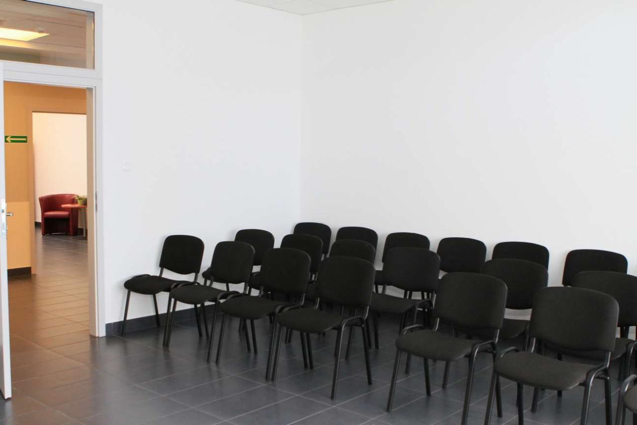 Poczekalnia z rzędem czarnych krzeseł na szarych płytkach, widoczne otwarte drzwi z zieloną strzałką i czerwonym fotelem w tle.