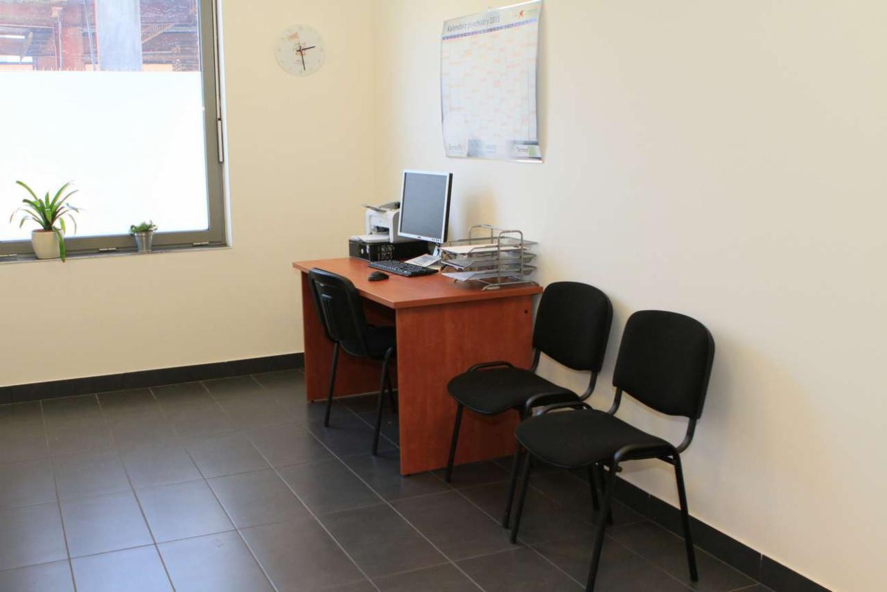 Wnętrze poczekalni: biurko z komputerem, drukarką i organizerem na dokumenty, dwie czarne krzesła dla pacjentów, kalendarz na ścianie, roślina doniczkowa na parapecie.