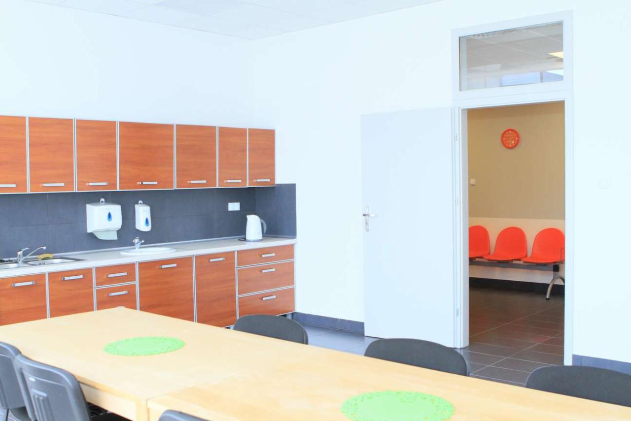 Wnętrze kliniki: kuchnia z drewnianymi szafkami i stołem, w tle poczekalnia z pomarańczowymi krzesłami i zegarem.