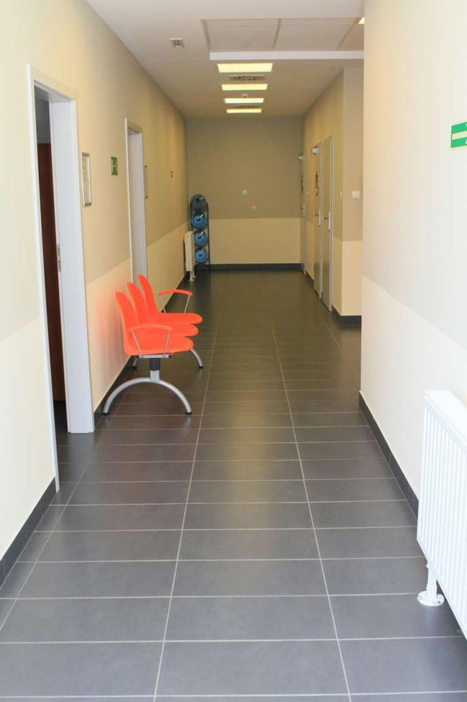 Długi korytarz w placówce medycznej z szarymi płytkami na podłodze, pomarańczowymi krzesłami dla pacjentów i białymi ścianami z beżowym pasem na dole.