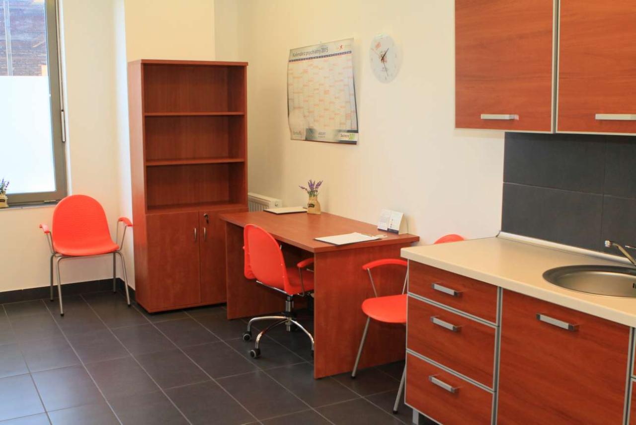 Wnętrze gabinetu z biurkiem, krzesłami, regałem i aneksem kuchennym w brązowej kolorystyce, kalendarz na ścianie.