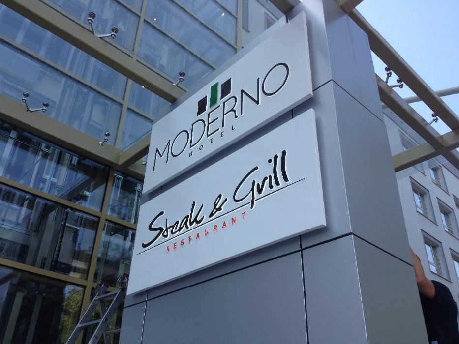 Zewnętrzny szyld reklamowy hotelu i restauracji z częściowo widoczną drabiną i przeszkloną fasadą budynku w tle.