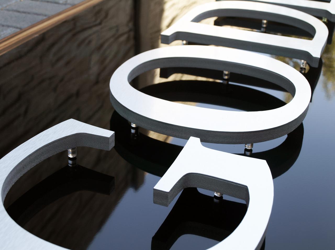 Szczegółowe zbliżenie na trójwymiarowe, srebrne litery z czarnym obramowaniem, zamontowane na błyszczącej, czarnej powierzchni, z widocznymi elementami montażowymi.