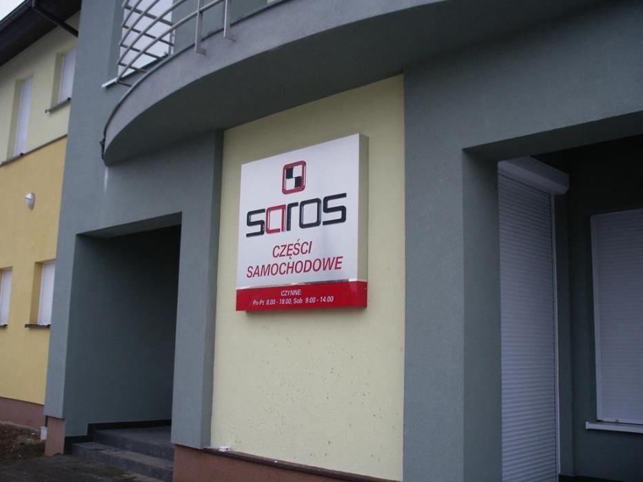 Szyld reklamowy firmy 'Saros Części Samochodowe' w Buku, z informacją o godzinach otwarcia, umieszczony na elewacji budynku o żółto-szarej kolorystyce.