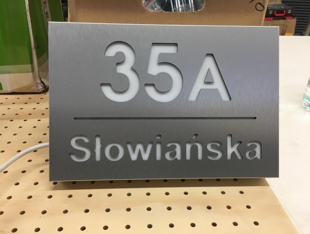 Metalowa tabliczka adresowa z numerem 35A i nazwą ulicy Słowiańska, wycięta laserowo, na drewnianym blacie.