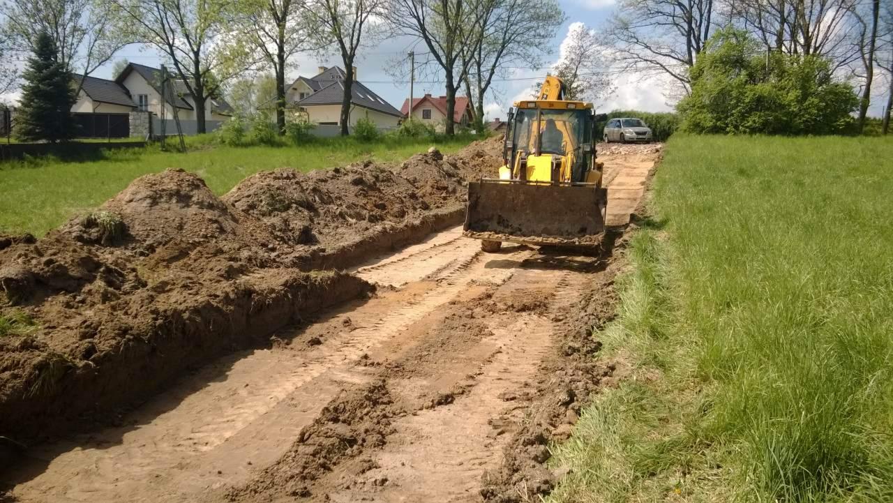 Żółta koparka podczas prac ziemnych na działce z widokiem na domy w tle, świeżo wykopany rów z pryzmami ziemi po bokach.