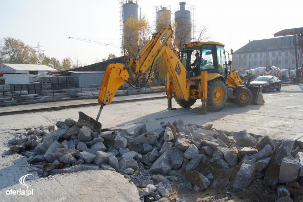 Żółta koparko-ładowarka z pneumatycznym młotem rozbija betonową nawierzchnię na placu budowy, w tle silosy i budynki.
