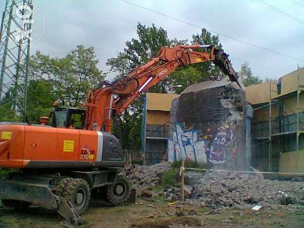 Pomarańczowa koparka Hitachi 210W z hydraulicznym młotem pneumatycznym kruszy betonowy element konstrukcyjny pokryty graffiti, otoczony gruzem i rusztowaniami.