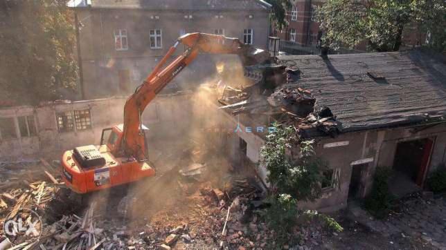 Pomarańczowa koparka z długim ramieniem rozbiera stary budynek z cegły i eternitu, unoszący się pył, w tle budynek mieszkalny.