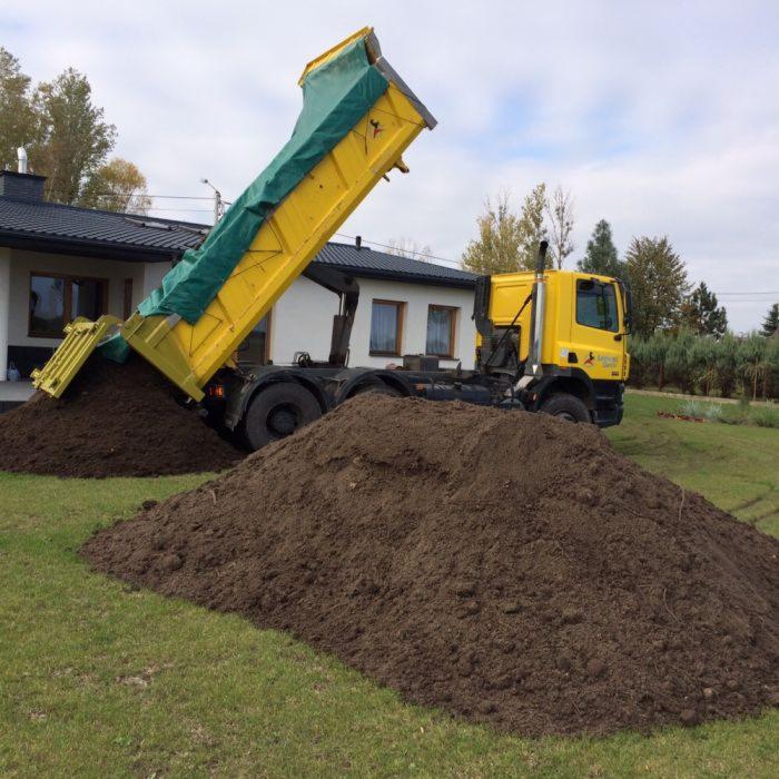 Żółta wywrotka z zieloną plandeką wysypuje ziemię na trawnik przed domem z białymi ścianami i ciemnym dachem.