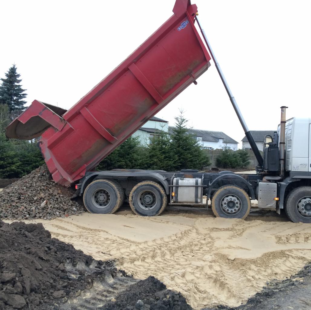 Ciężarówka wywrotka z podniesioną czerwoną skrzynią, wysypująca gruz na piaszczysty teren budowy, widoczne ślady opon i drzewa w tle.