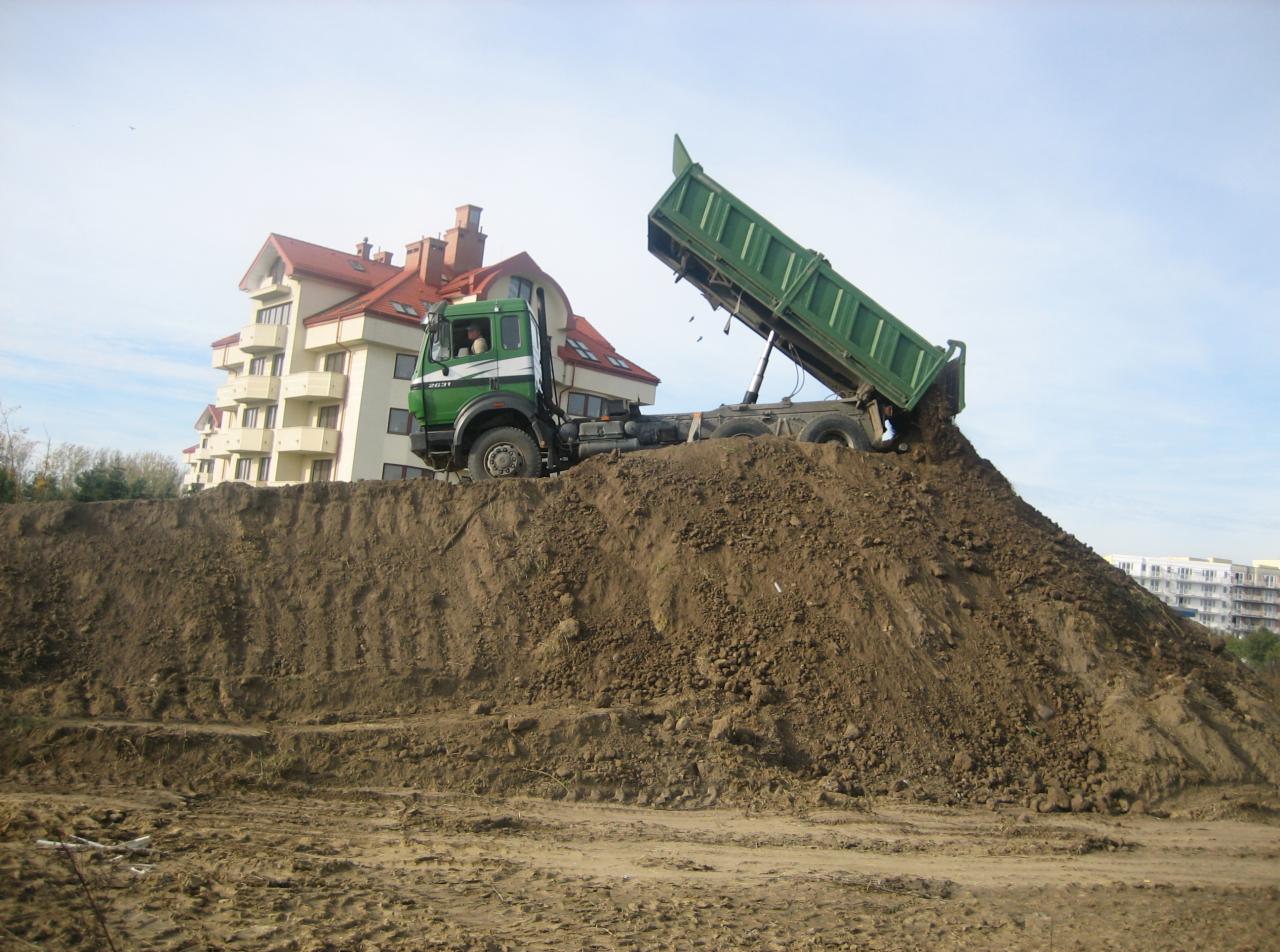 Zielona wywrotka wysypuje ziemię na pryzmę, w tle budynek mieszkalny.
