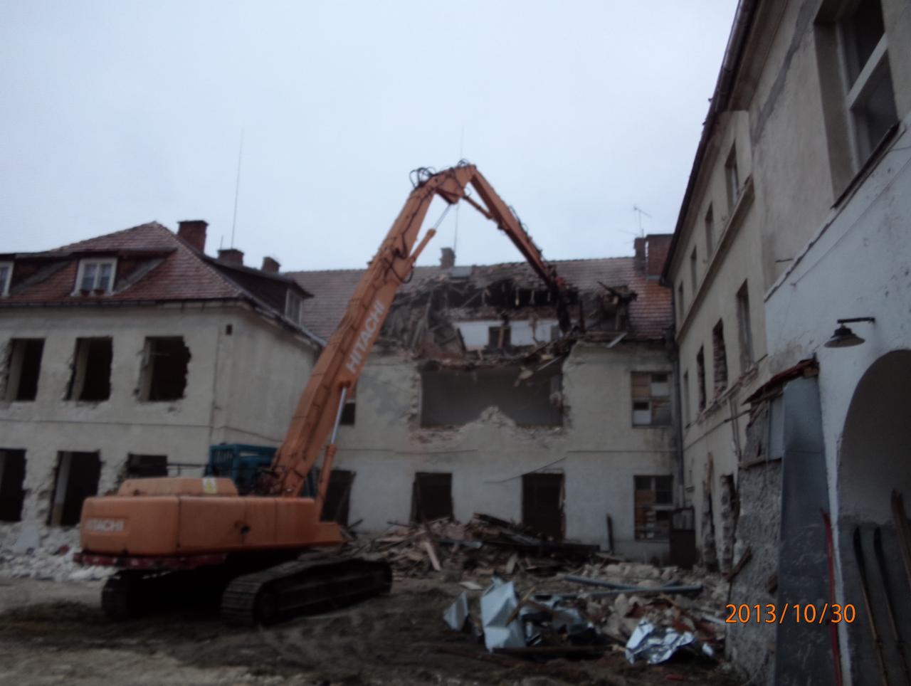 Pomarańczowa koparka Hitachi podczas rozbiórki budynku z czerwonym dachem, gruz leży na ziemi, pochmurne niebo.