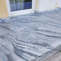 Zewnętrzny parapet okienny z szarego kamienia z wyraźnym, falistym wzorem, zamontowany przy oknie w jasnej ramie, z fragmentem żółtej i białej ściany w tle.