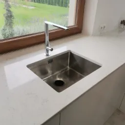 Biały blat kuchenny z szarymi żyłkami, zintegrowany zlewozmywak ze stali nierdzewnej i nowoczesna bateria, widok na ogród przez drewniane okno.