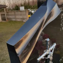 Nowoczesny, czarny pomnik z elementami piaskowanymi i srebrnym krucyfiksem, zlokalizowany na cmentarzu w Popowie, na tle drzew bez liści.