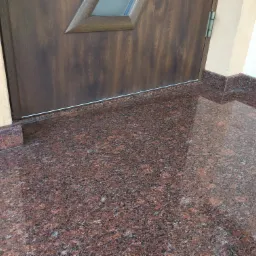 Błyszczący, czerwono-brązowy kamienny taras przed wejściem do domu z drewnianymi drzwiami z szybą, wykończony kamiennymi obrzeżami.