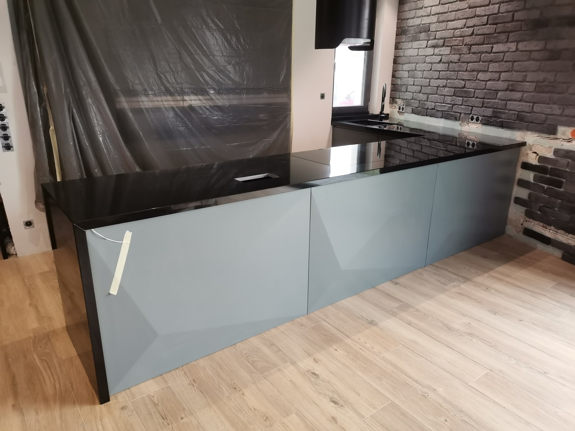 Czarny blat kuchenny z szarymi frontami szafek, częściowo zabezpieczony folią malarską, widoczny fragment ściany z cegły i drewniana podłoga w trakcie montażu.