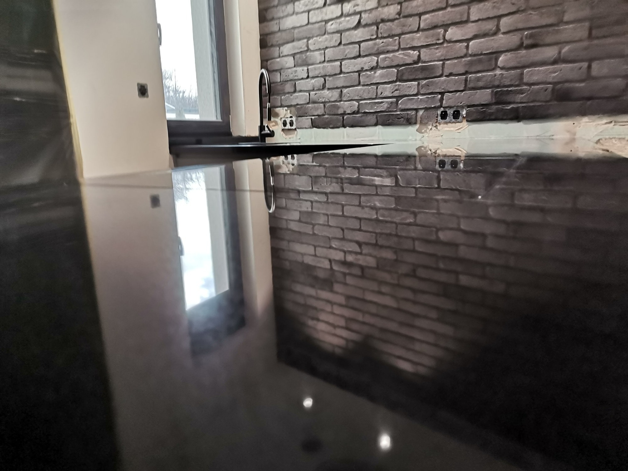 Błyszczący, czarny blat w kuchni, z widoczną armaturą i ceglaną ścianą w tle, odbijającą się w powierzchni blatu.