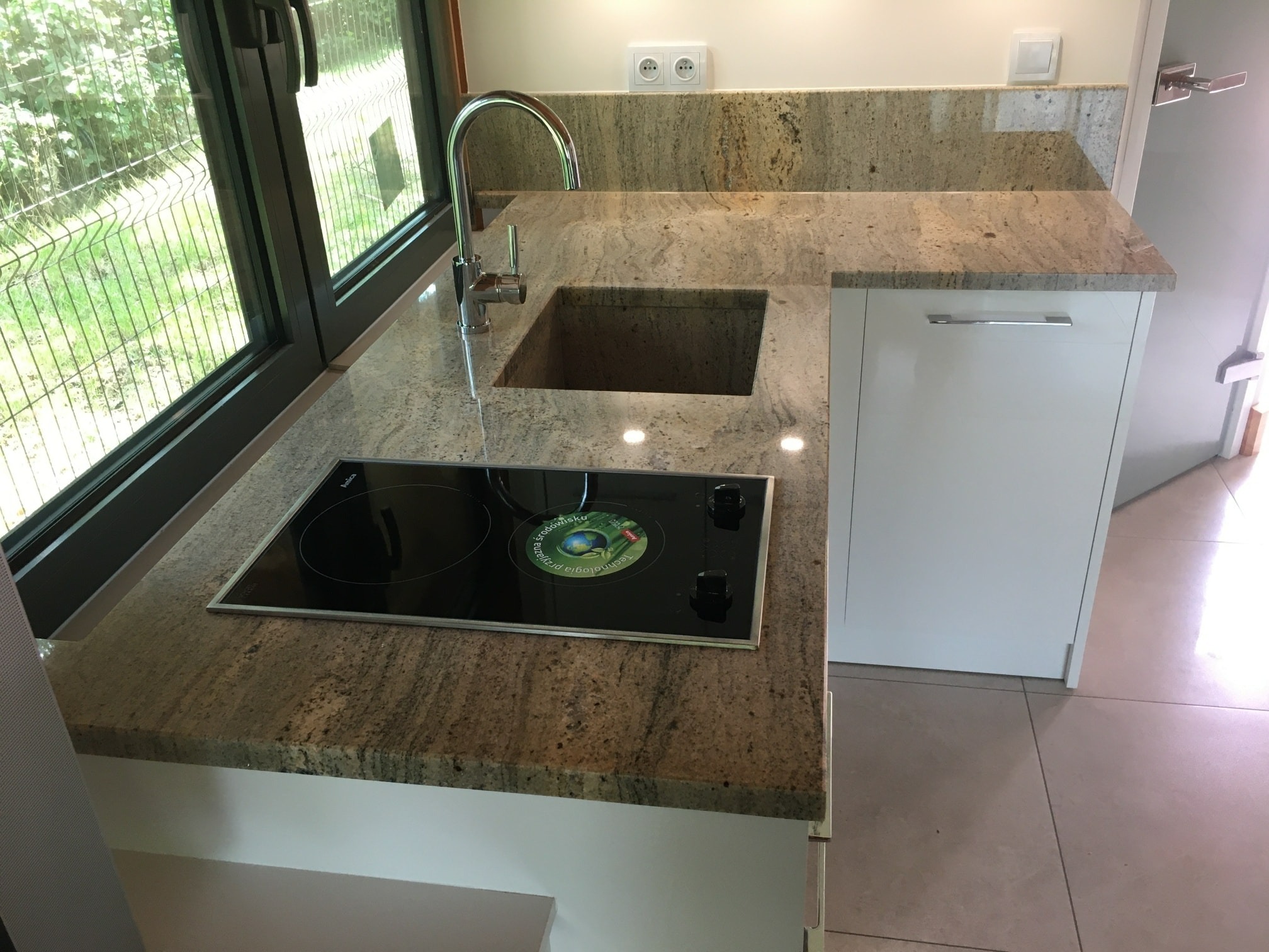 Kuchenny blat z kamienia o brązowo-beżowej teksturze, z wbudowanym zlewem i płytą indukcyjną, widok z góry na fragment zabudowy kuchennej przy oknie.