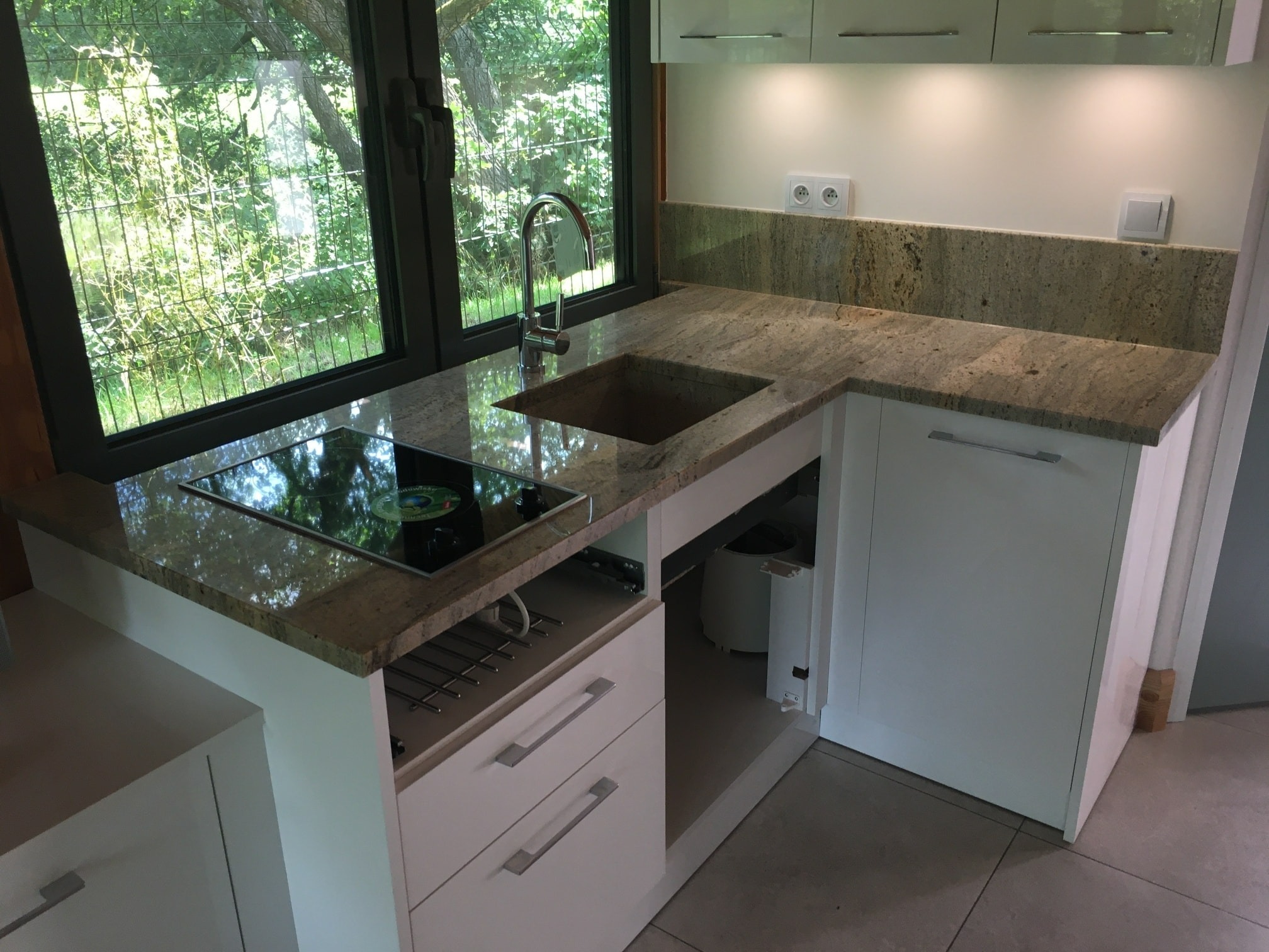 Kuchnia z blatem z kamienia naturalnego, zlew i płyta indukcyjna wbudowane w blat, białe szafki, widok na zieleń za oknem.