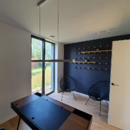 Nowoczesne wnętrze biurowe z granatową ścianą, drewnianymi półkami i minimalistycznymi krzesłami, uzupełnione biurkiem z czarnym blatem i drewnianymi elementami oraz oświetleniem liniowym.