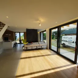 Jasne, przestronne wnętrze domu z drewnianą podłogą, schodami z białymi barierkami, minimalistycznym kominkiem, szarą sofą, czarnym panelem dekoracyjnym na ścianie i widokiem na zewnątrz przez...