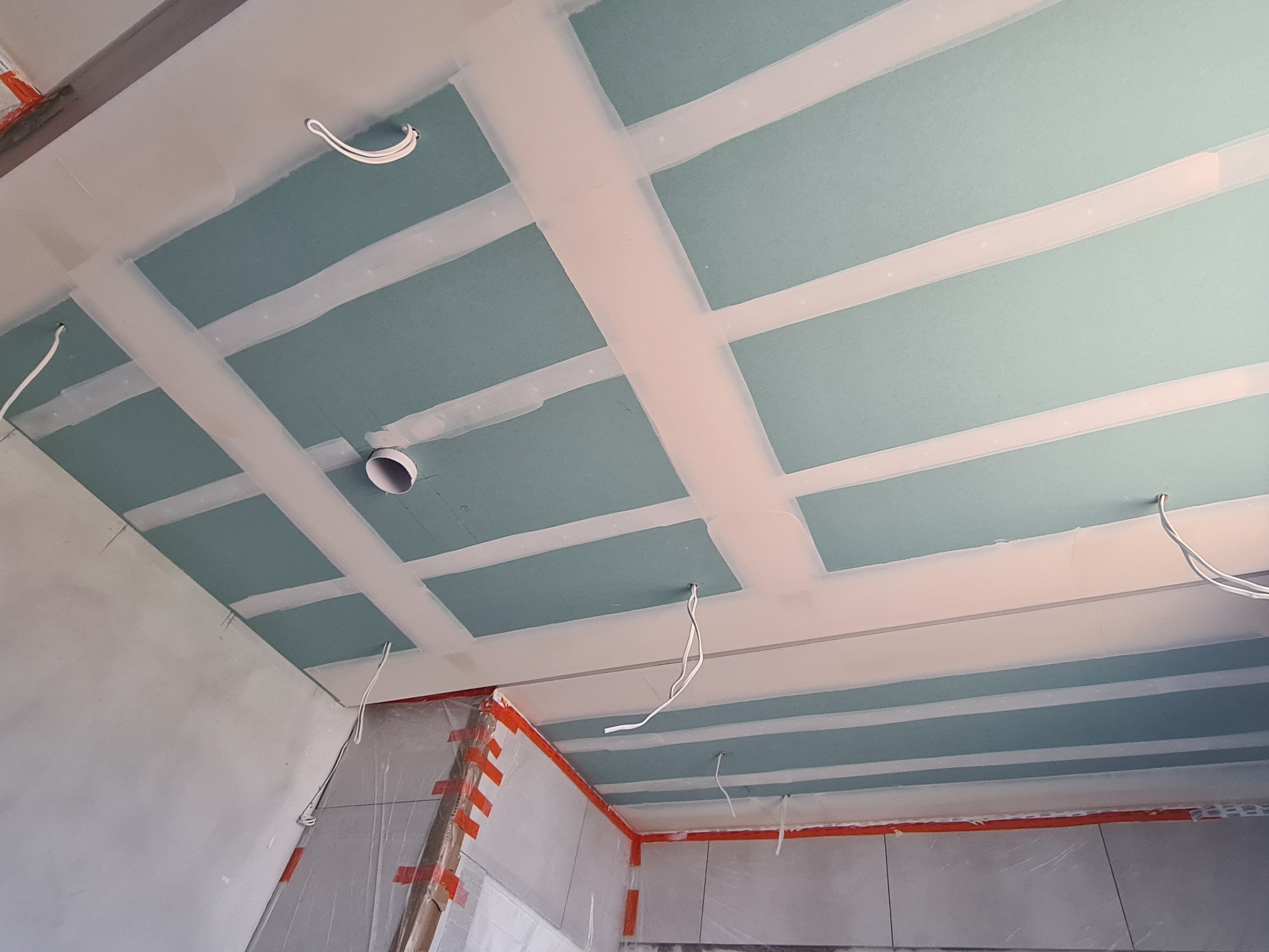 Sufit z zamontowanymi płytami gipsowo-kartonowymi, widoczne łączenia wzmocnione taśmą, wystające przewody elektryczne i otwór wentylacyjny, zabezpieczone folią ściany w tle.