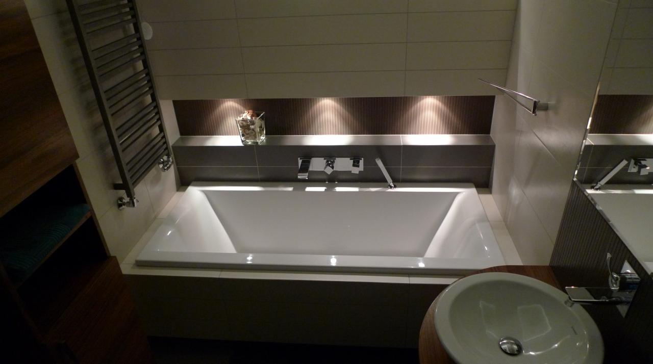 Nowoczesna łazienka z białą wanną w zabudowie, podświetloną półką z dekoracją i chromowaną armaturą, obok umywalka i grzejnik drabinkowy.