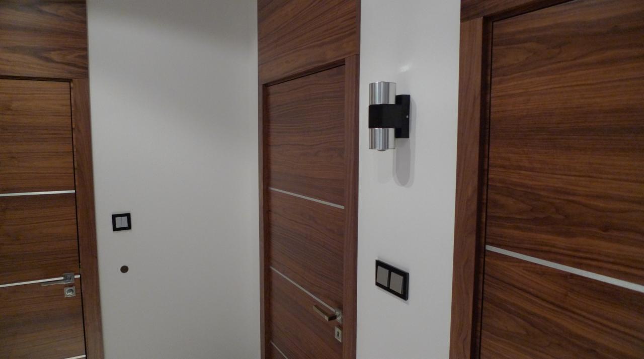 Nowoczesne, drewniane drzwi wewnętrzne z poziomymi, metalowymi akcentami, oświetlenie ścienne w postaci dwóch srebrnych cylindrów na czarnym tle, minimalistyczny włącznik światła i gniazdko...