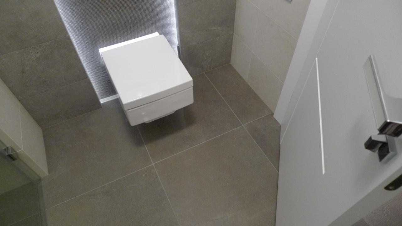 Nowoczesna toaleta podwieszana z podświetleniem LED w zabudowie, szare płytki podłogowe i ścienne, białe drzwi z chromowanym uchwytem.