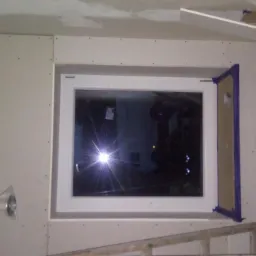 Montaż płyty karton gips wokół okna, widoczne łączenia płyt i zabezpieczone taśmą malarską obramowanie okna, fragment drabiny i rolka materiału na pierwszym planie.