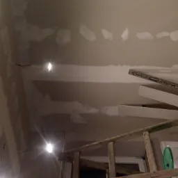 Nisko kątowe ujęcie sufitu w trakcie prac wykończeniowych; widoczne ślady po szpachlowaniu, wystające przewody elektryczne, drabina drewniana z narzędziami i materiałami budowlanymi w tle.