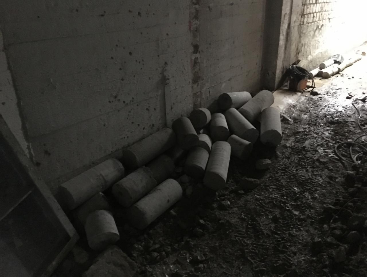 Rdzenie betonu ułożone na ziemi w ciemnym pomieszczeniu, widoczne ślady opon i sprzęt w tle.