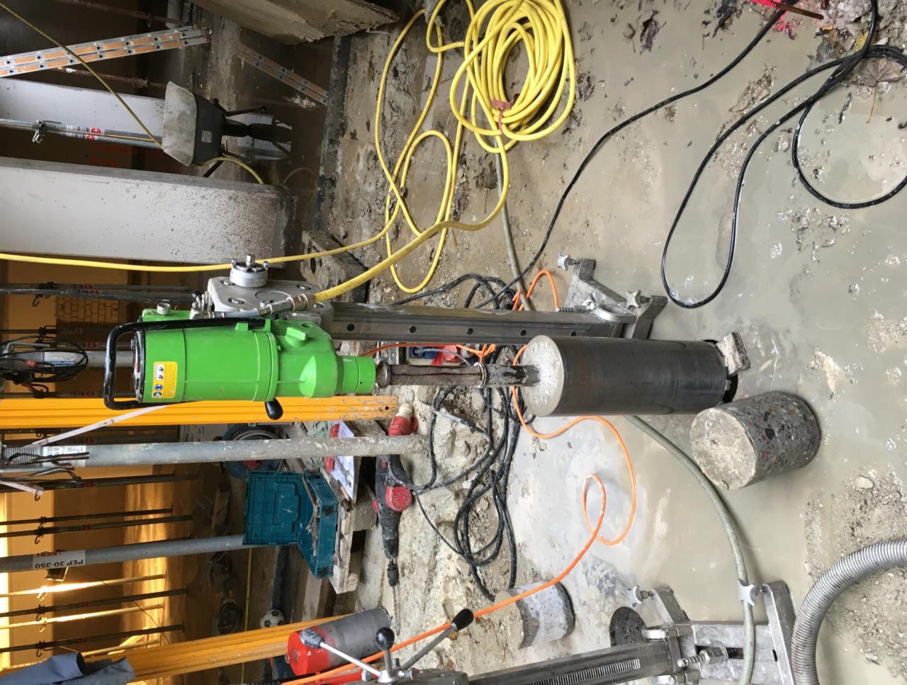 Wiertnica rdzeniowa w trakcie pracy; zielona maszyna wiercąca zamocowana na metalowej konstrukcji, obok wywiercony rdzeń betonowy, woda na ziemi.