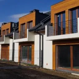 Szereg nowych domów z drewnianą elewacją i małymi balkonami, jeszcze w trakcie wykończenia, ogrodzone siatką.