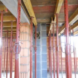 Wnętrze w trakcie budowy: widok na strop podparty czerwonymi stemplami budowlanymi, ściany z cegły ceramicznej dziurawki i bloczków betonowych, widoczne drewniane elementy szalunku stropu.