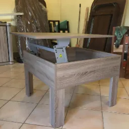 Rozłożony stolik kawowy z mechanizmem podnoszenia blatu, wykończony laminatem imitującym jasne drewno, widoczne elementy montażowe i tło z wyposażeniem pokoju.