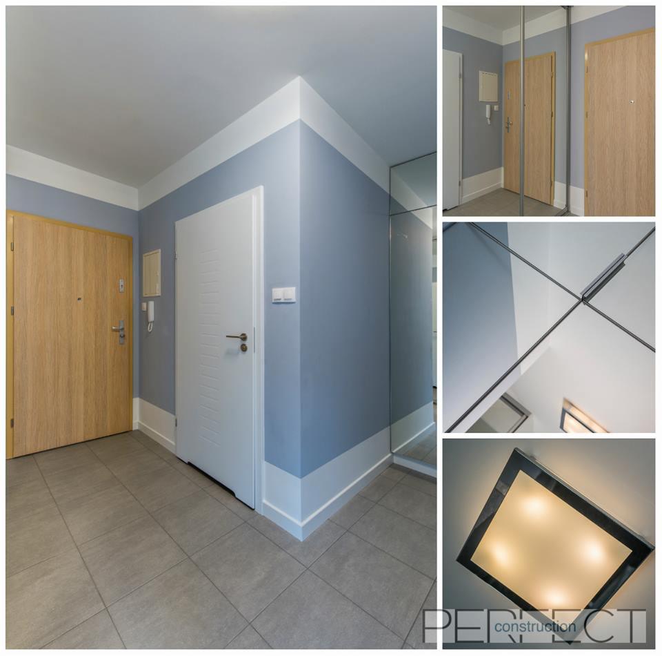 Nowoczesny przedpokój w odcieniach szarości i błękitu z drewnianymi drzwiami wejściowymi, białymi drzwiami wewnętrznymi, lustrem na ścianie i kwadratową lampą sufitową.