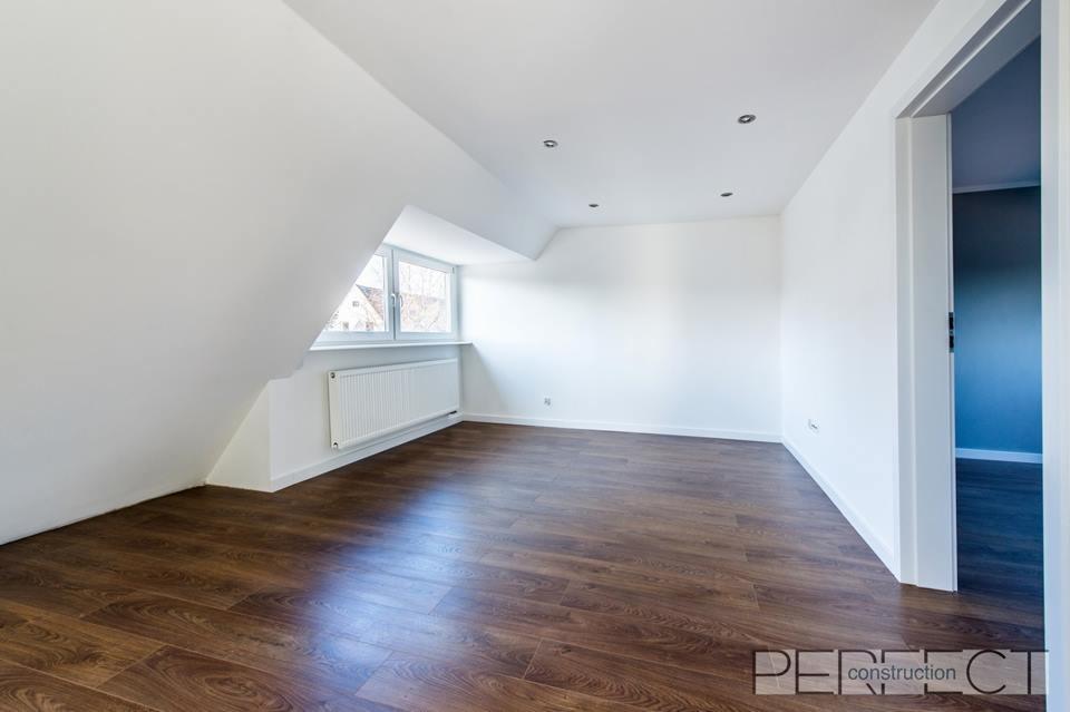 Poddasze po remoncie z nową, ciemną podłogą imitującą drewno, białymi ścianami, grzejnikiem pod oknem i otwartymi drzwiami do sąsiedniego pomieszczenia.