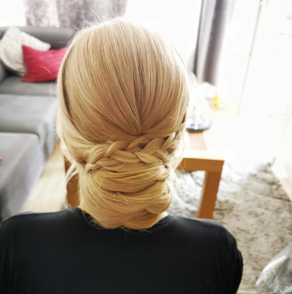 Upięcie blond włosów z warkoczem z tyłu głowy, widoczne z perspektywy osoby siedzącej na krześle, w tle szara kanapa i drewniany stolik.