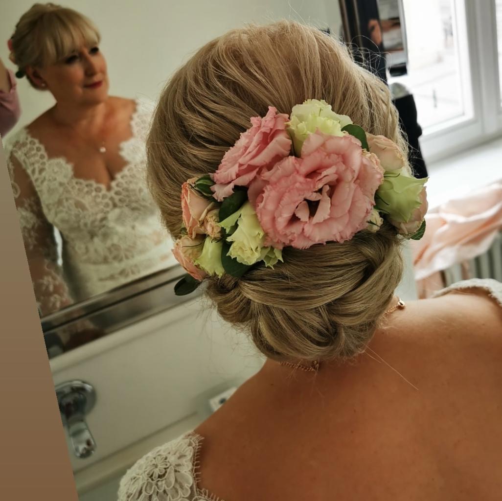 Upięcie ślubne z blond włosów z wpiętymi różowymi i kremowymi kwiatami, widok z tyłu, odbicie w lustrze ukazuje kobietę w koronkowej sukni.