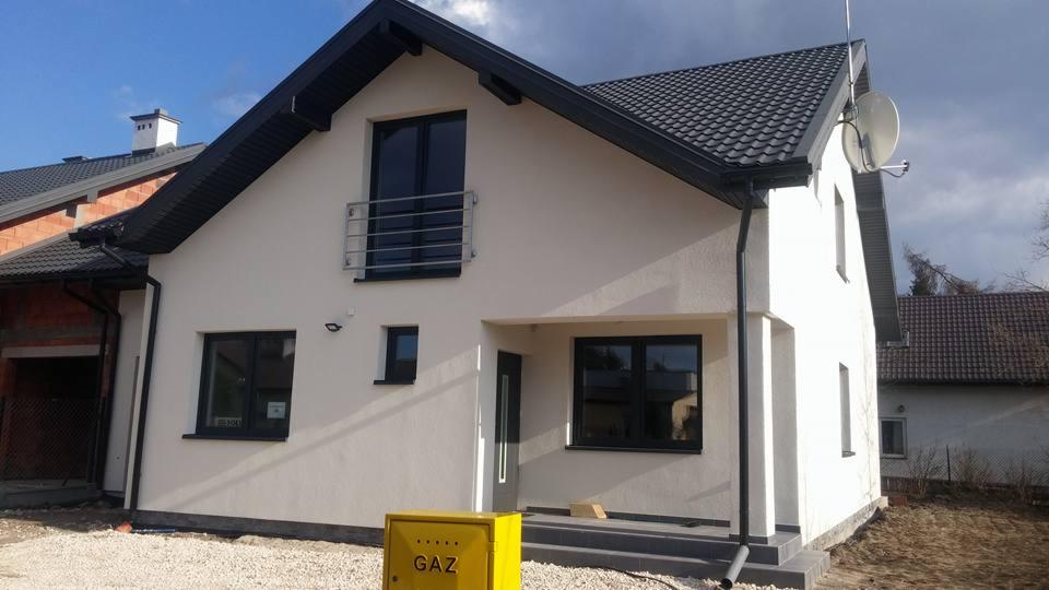 Nowoczesny dom jednorodzinny z białym tynkiem, ciemną stolarką okienną i dachem z dachówki, widoczna skrzynka gazowa w kolorze żółtym i niezakończona elewacja sąsiedniego budynku.