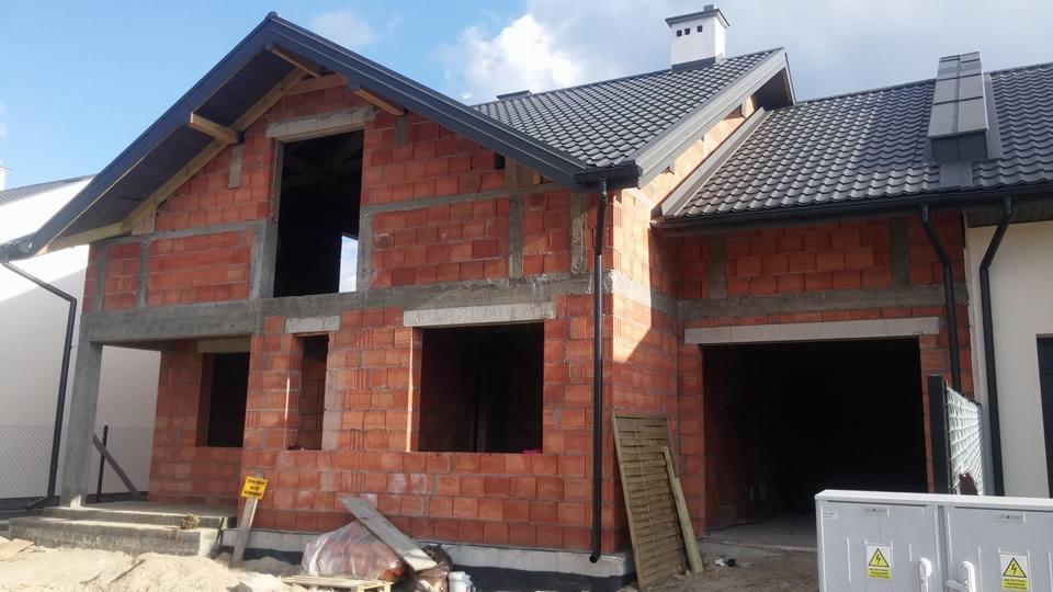 Surowy stan budynku z czerwonej cegły z widocznymi otworami okiennymi i drzwiowymi, czarnym dachem z dachówki i elementami szalunku.